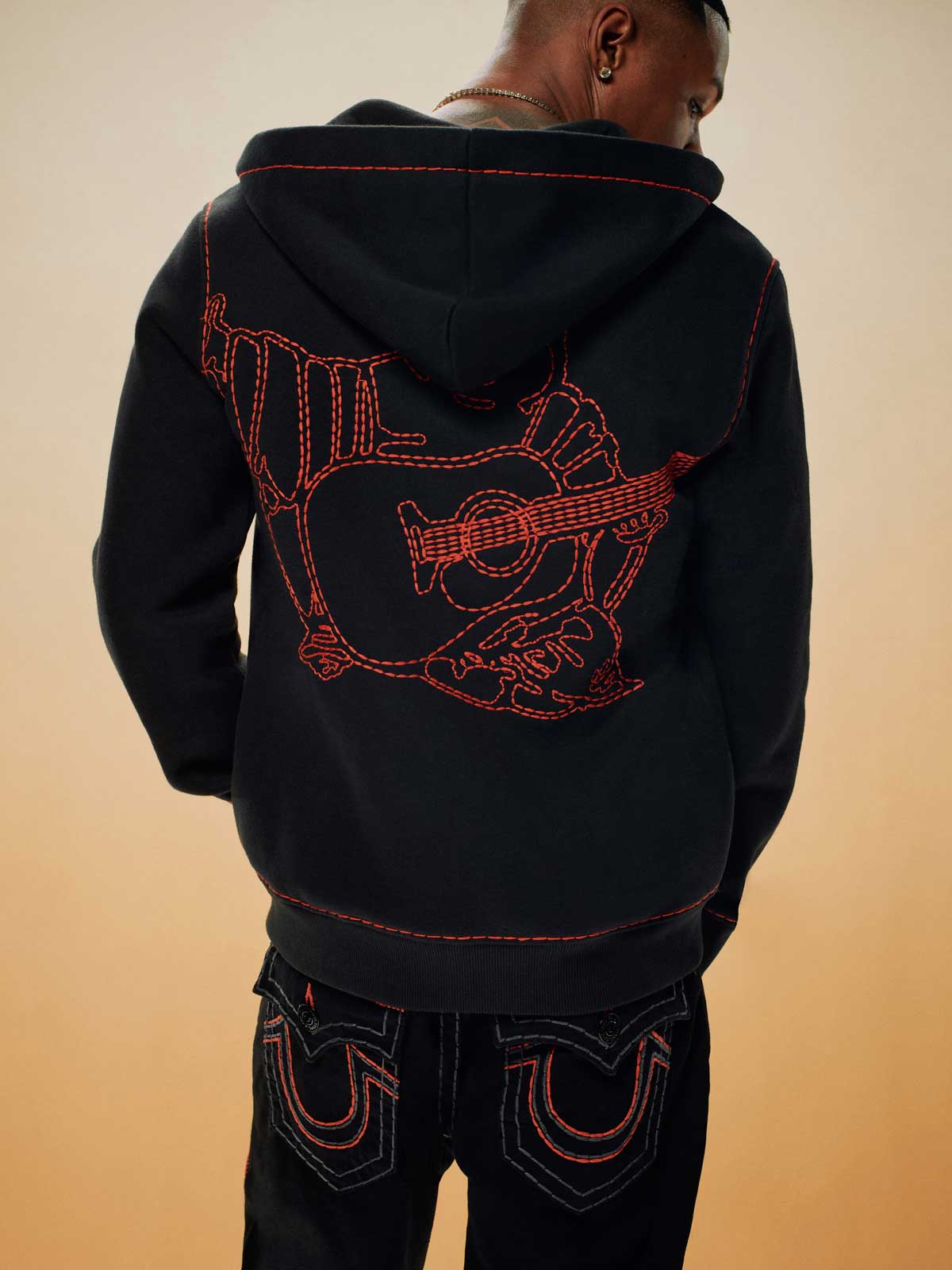 Man wears black true religion hoodie with red stitching of a guitar, and black true religion pants with red horseshoe stitching