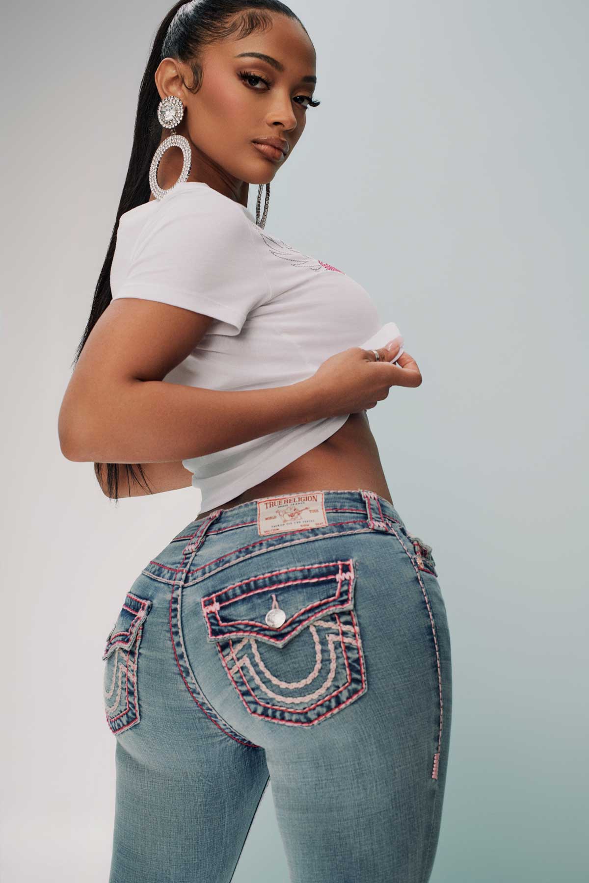 Woman wears true religion jeans with pink and white stitching on the back, a white baby tee, and hoops