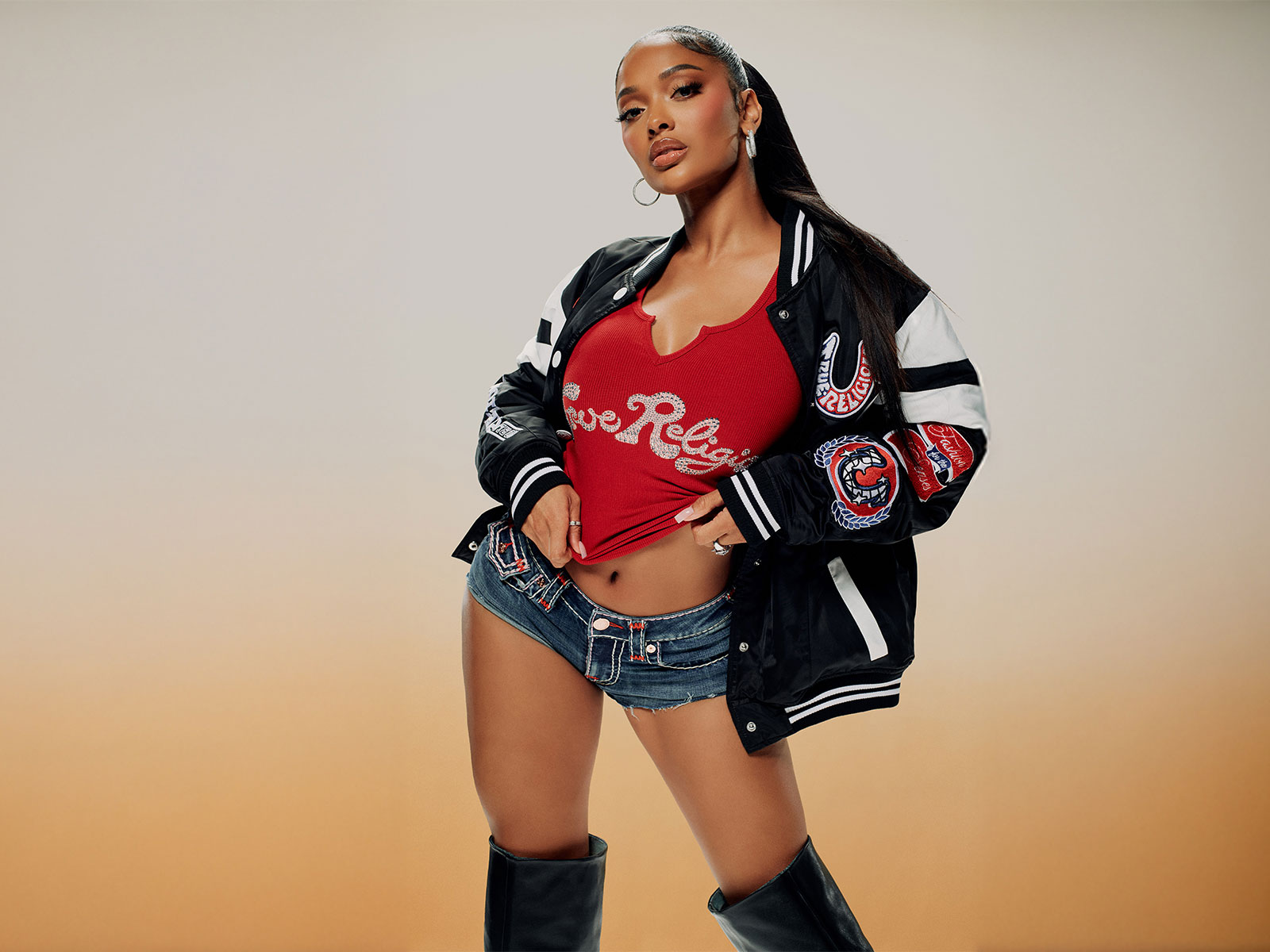 Woman wears true religion low rise jean shorts, a true religion red tee shirt, and a true religion black, white, and red varsity jacket 