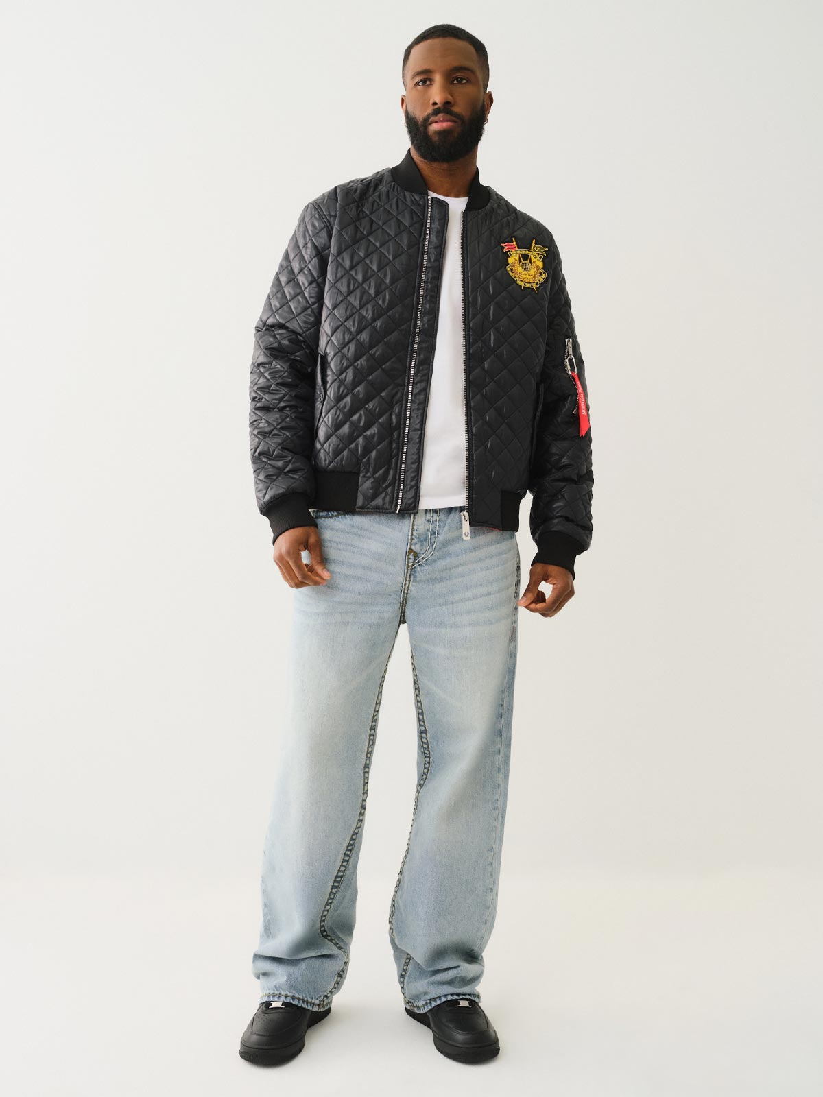 Man wears a true religion black quilted bomber jacket and true religion light wash baggy jeans