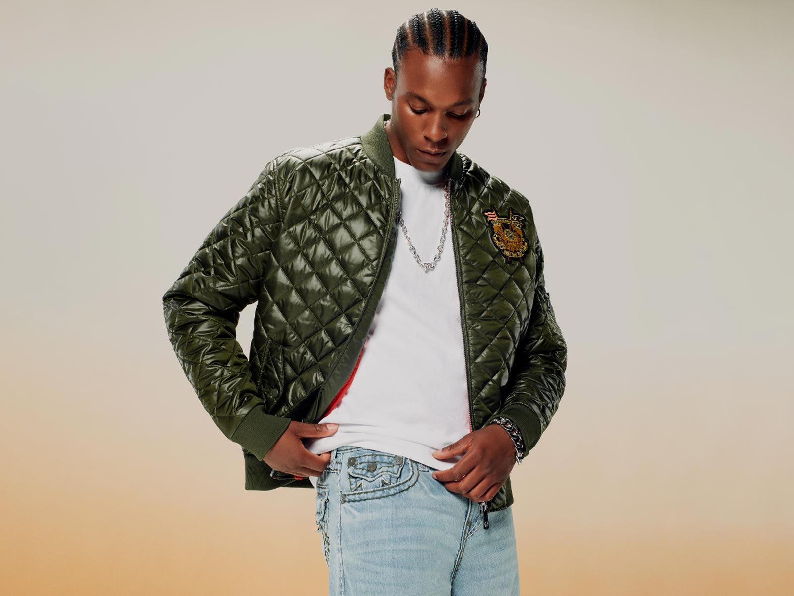 Man wears a true religion quilted green bomber jacket, a white tee, and true religion light wash jeans 
