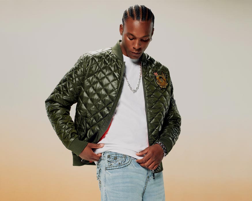 Man wears a true religion quilted green bomber jacket, a white tee, and true religion light wash jeans 
